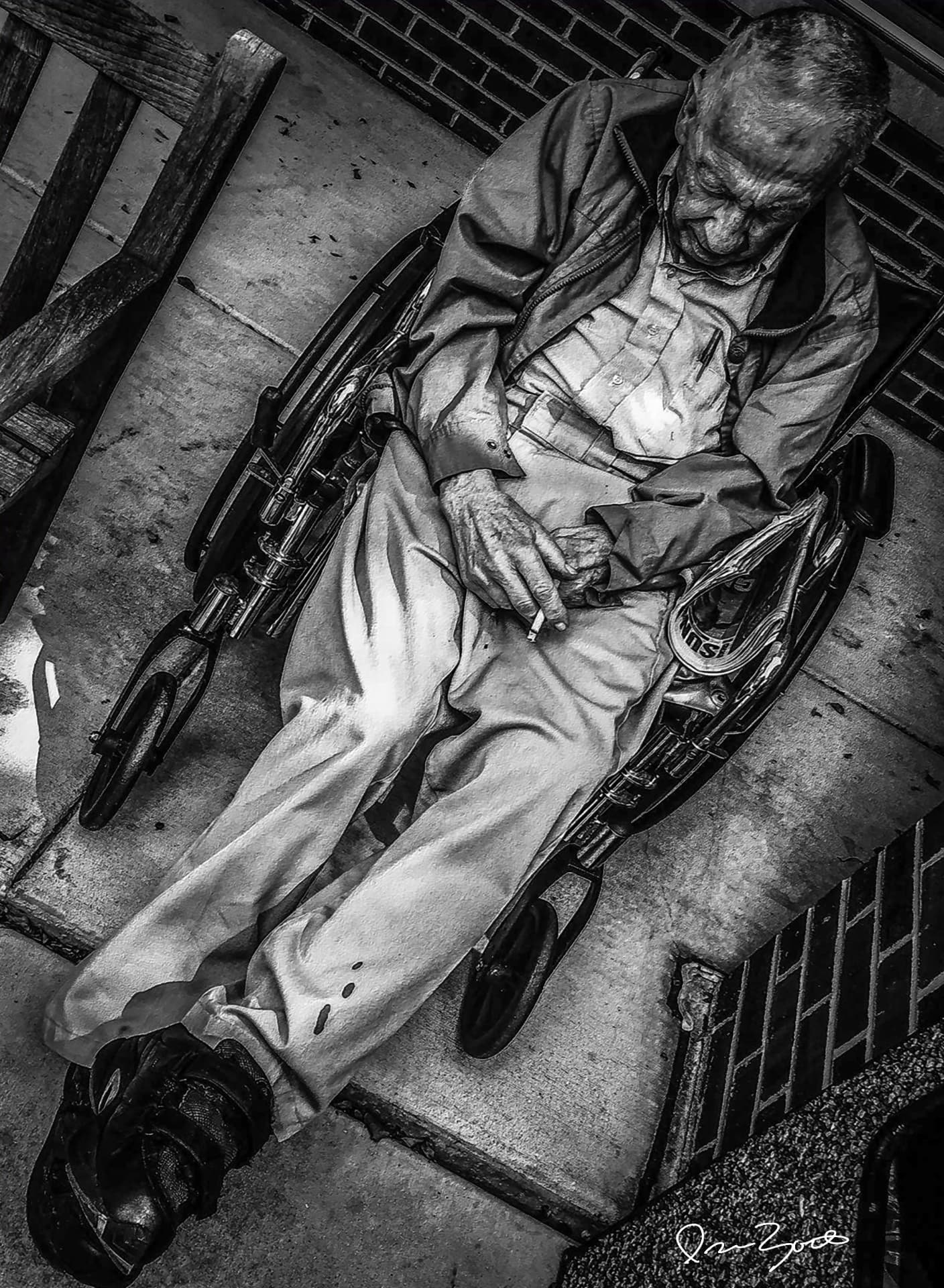 Black and white elevated photograph of an elderly man in a wheelchair on a concrete stoop holding a cigarette, brick wall behind.