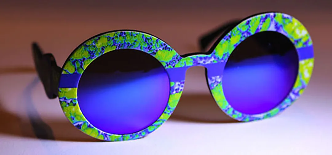 Round sunglasses with green and violet patterned frames and blue mirrored lenses on a warm background.